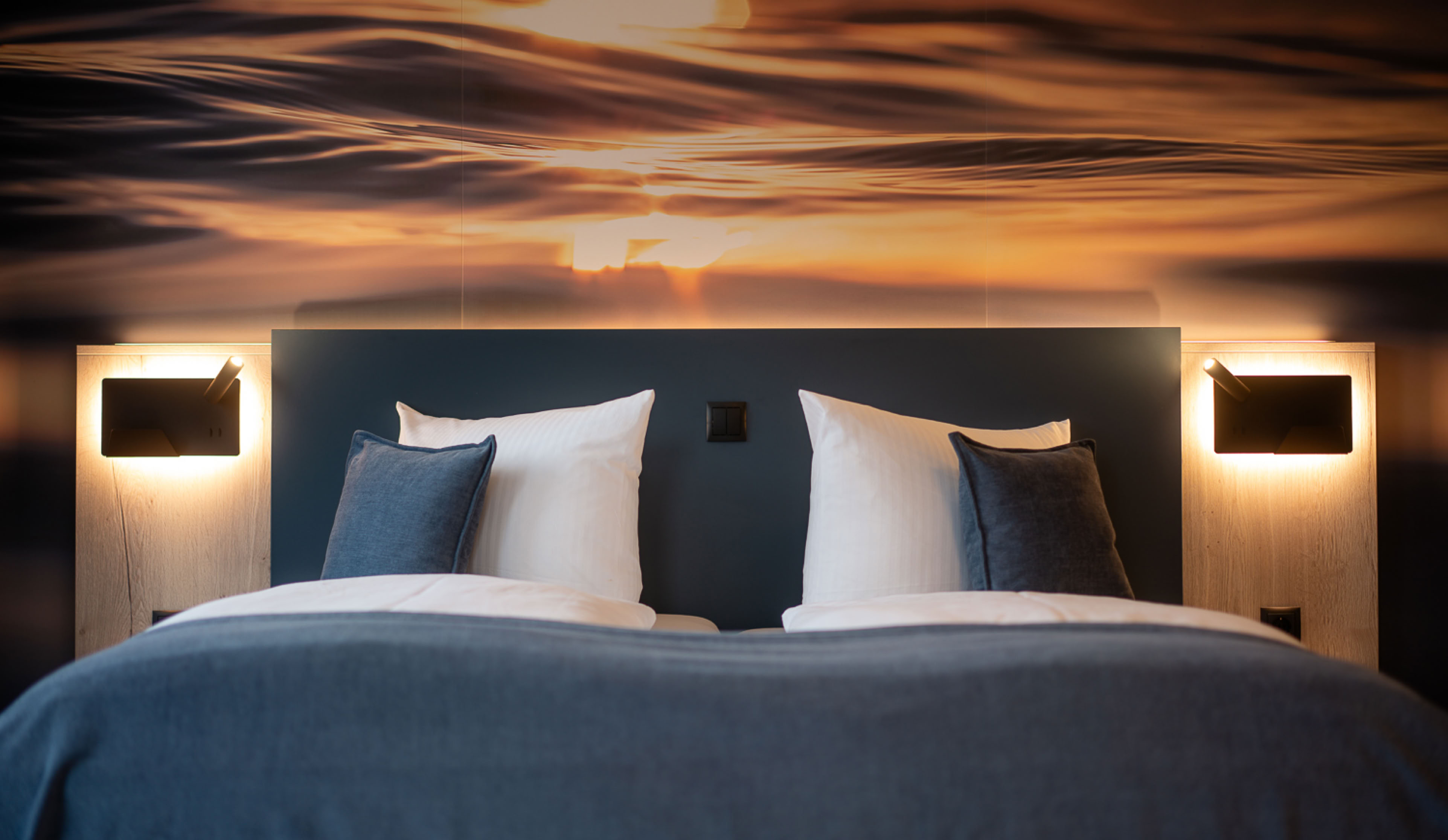 Ein Doppelbett mit blauer Bettdecke und weißen Kissen, flankiert von zwei Wandleuchten, vor einer Wand mit einem abstrakten Muster in warmen Farben.